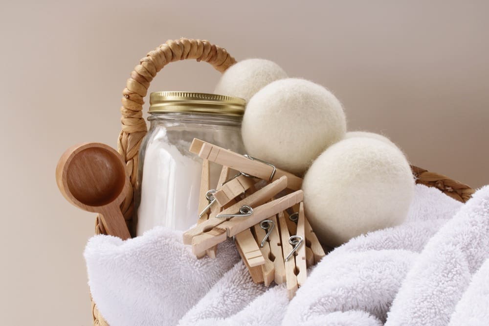 A basket containing a glass jar with a gold lid, three wool dryer balls, several wooden clothespins, a wooden scoop, and a folded white towel.