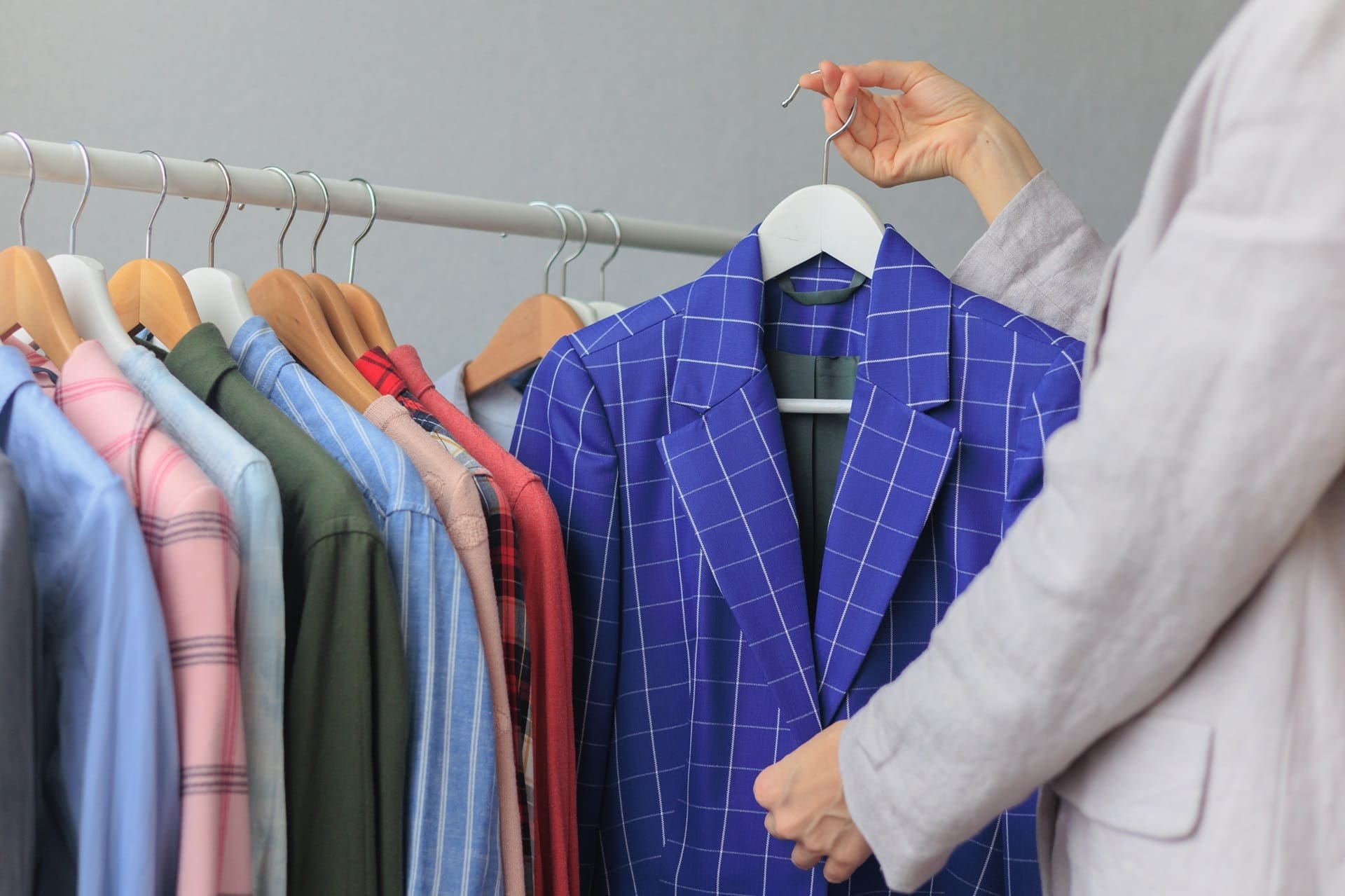 A person selects a blue checkered blazer from a clothing rack filled with various shirts and jackets on wooden hangers.