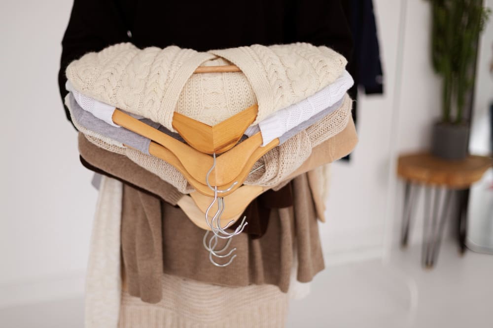 Person holding a stack of knit sweaters on wooden hangers, with several empty hangers, in a bright room with minimal decor.