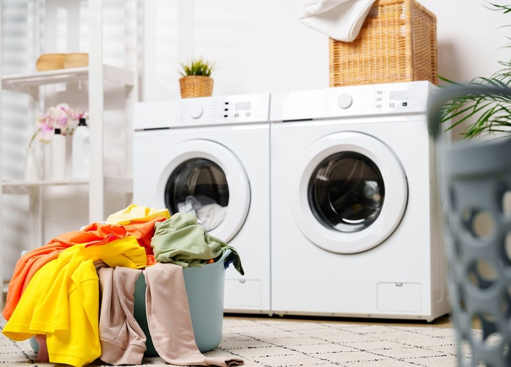 A laundry room with a washer and dryer, a basket overflowing with colorful clothes in the foreground, and shelves and a wicker basket in the background.