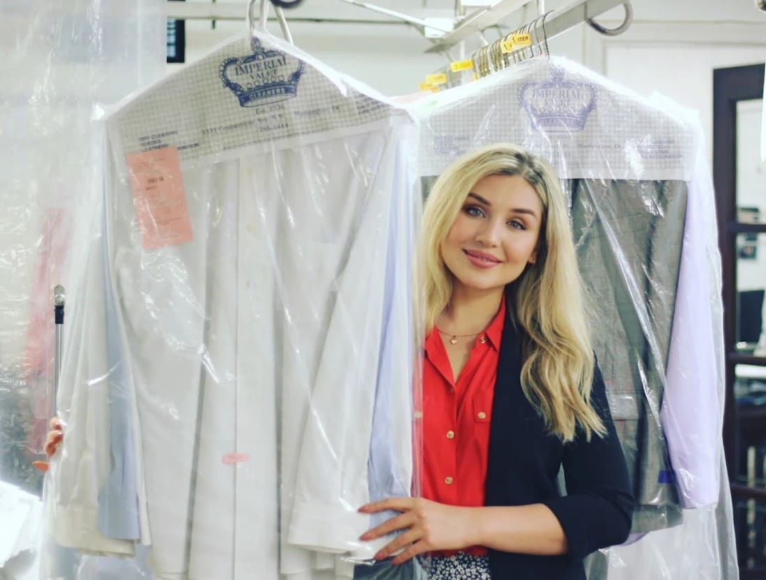 A woman stands in a dry cleaning shop, smiling and holding freshly cleaned clothes covered in plastic garment bags on hangers.